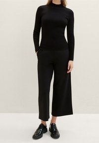 Top lungo a maniche nere a coste con colletto alto, abbinato a pantaloni neri a gamba larga. Texture liscia e silhouette aderente. Scarpe nere con lacci completano il look.