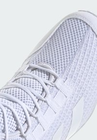 Zapatilla deportiva blanca con un upper de malla texturizada, cordones planos y acentos sintéticos lisos. Logotipo de tres rayas distintivo en el costado.