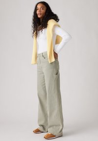 Γυναίκα με μακριά, σγουρά μαλλιά, που φοράει λευκή μπλούζα με μακριά μανίκια, κίτρινο πλεκτό πουλόβερ στους ώμους, άνετο ανοιχτό γκρι παντελόνι και καφέ αθλητικά παπούτσια.