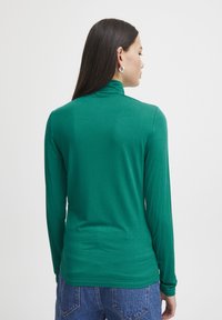 Langarm-Grüntop aus weichem Stoff mit hohem Kragen. Hat einen schmalen Schnitt und eine glatte Textur, kombiniert mit hochtaillierten blauen Jeans.