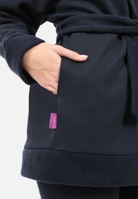 Marineblaue Fleece-Robe mit strukturierten Bündchen, einem Taillengürtel, einer Seitentasche und einem kleinen lila Etikett, das am Saum angenäht ist. Weiches und gemütliches Material.