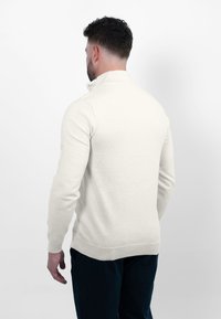 Lichtbeige gebreide trui met een hoge kraag, ribgebreide manchetten en zoom, aansluitende silhouet, gladde textuur, gedragen over donkerblauwe broek.