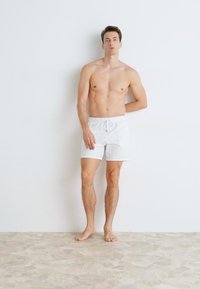 Bela kopalna šortsa iz lahkega materiala, z elastičnim pasom in vrvico za zategovanje. Prikazana na moškem modelu, ki stoji bos.