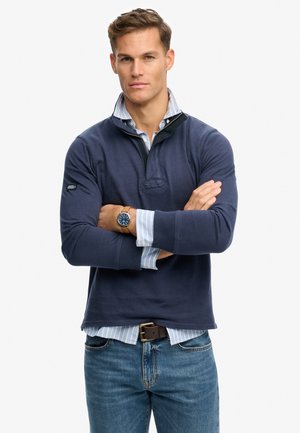 Mężczyzna ze skrzyżowanymi ramionami, ubrany w granatowy sweter, niebieską koszulę w paski, niebieskie dżinsy i zegarek na rękę, stojący na jednolitym białym tle.