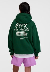Green hoodie with white text and graphics on the back, featuring “BEE’S STADIUM RESTAURANT” and “hummel” logo, paired with pink shorts.