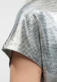 Nærbillede af en bar arm iført en kortærmet top i metallisk sølvfarvet tekstureret stof med synlige sømme ved ærmelinningen.