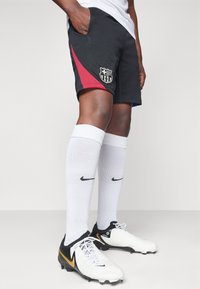 Svarta sportshorts med röd sidpanel, med en logotyp; tillsammans med vita knähöga strumpor och svart/vita fotbollsskor.