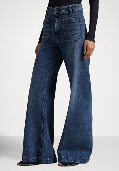 Γυναίκα που φοράει μπλε σκουρό τζιν παντελόνι υψηλής μέσης με φαρδύ πόδι, μπροστινές τσέπες με επιθέματα και μαύρα παπούτσια, στέκεται με ένα χέρι χαλαρό δίπλα της.