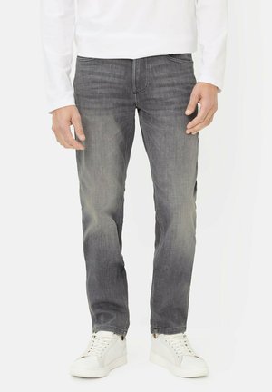 Mann trägt ausgewaschene graue Jeans, weißes langärmliges Hemd und weiße Turnschuhe und steht vor einem weißen Hintergrund.