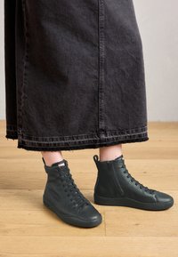 Personne portant des baskets en cuir montantes noires avec une fermeture éclair sur le côté et des lacets, se tenant sur un parquet, sous une jupe sombre de longueur mi-cuisse.