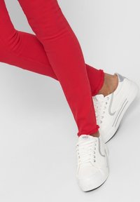 Jeans skinny rojos con dobladillos deshilachados, combinados con zapatillas blancas que tienen un acento plateado y un diseño texturizado. Tejidos suaves, silueta ajustada.