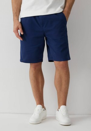 Mężczyzna ubrany w granatowe szorty, białe trampki i białą koszulę, stojący z jedną ręką w kieszeni na jednolitym tle.