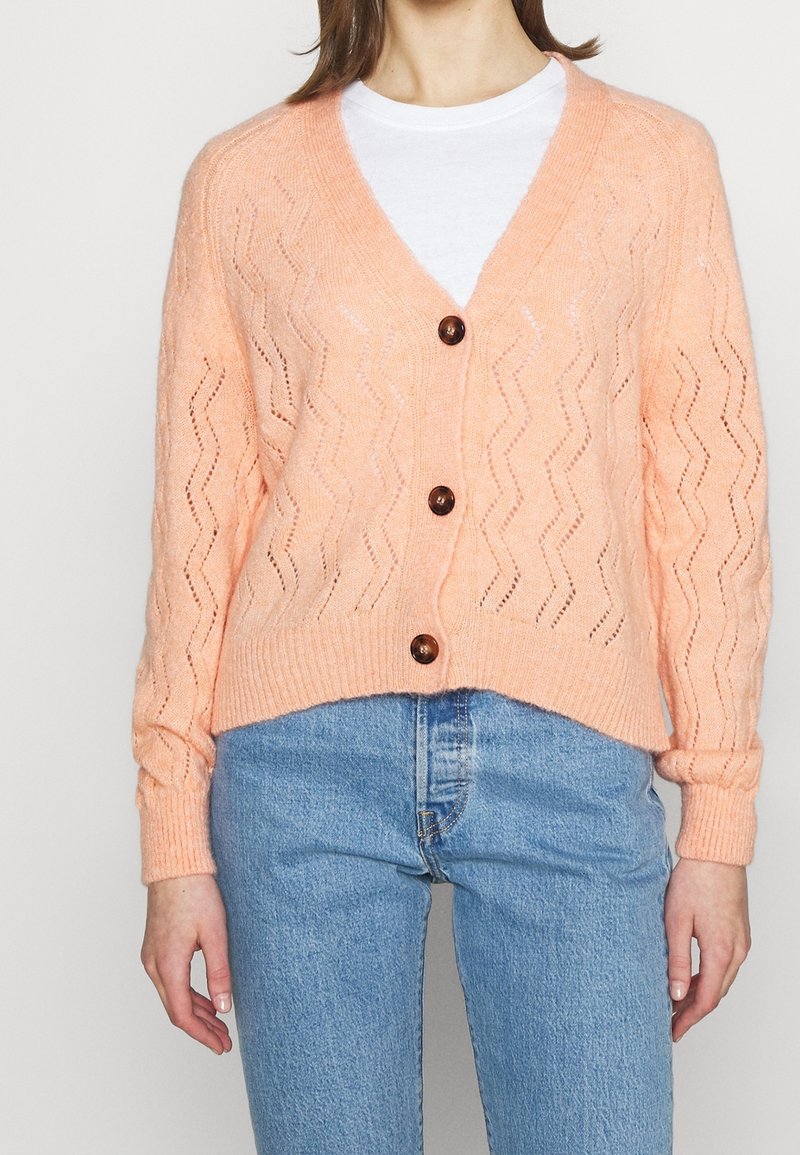 Cardigan en maille pêche avec col en V, poignets côtelés et fermeture à boutons. Présente un motif en zigzag et une coupe courte, porté avec un jean clair.