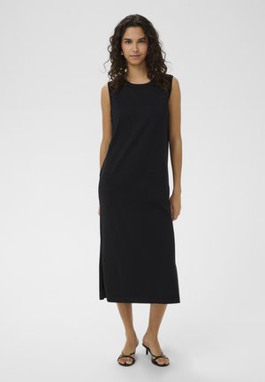 Kobieta o kręconych włosach, ubrana w czarną, bezrękawową sukienkę midi i czarne sandały na obcasie, stojąca na jednolitym jasnym tle.