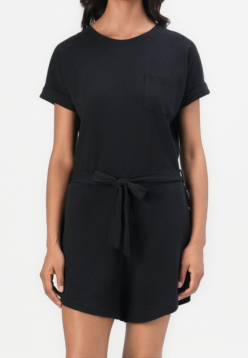 Femme portant une combinaison-short noire à manches courtes avec une poche avant et une ceinture en tissu nouée à la taille, debout devant un fond blanc.