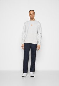 Gray sweatshirt with orange logo, paired with navy pants. Model wears white sneakers. Sleek design, casual fit, and soft texture.