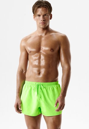 Muskularny mężczyzna z mokrą skórą, ubrany w jaskrawozielone kąpielówki, stojący na jednolitym jasnym tle.