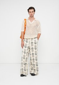 Un joven está de pie vistiendo una camiseta corta de malla blanca, pantalones anchos color crema con estampados florales negros, zapatos negros y un bolso naranja de hombro.