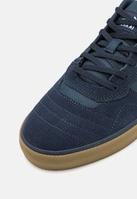 Marineblauer Wildledersneaker mit einem strukturierten Mesh-Bereich, runder Spitze, Gummisohle und auffälligen Nähten. Die Schnürsenkel sind ebenfalls marineblau.