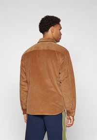 Brown corduroy shirt with a collar, featuring a textured surface and long sleeves. The back has a curved hem and a relaxed fit.