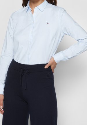 Kvinde iført lyseblå skjorte med knapper og et lille logo på brystet samt mørke bukser med snøre og diskret broderi på hoften.