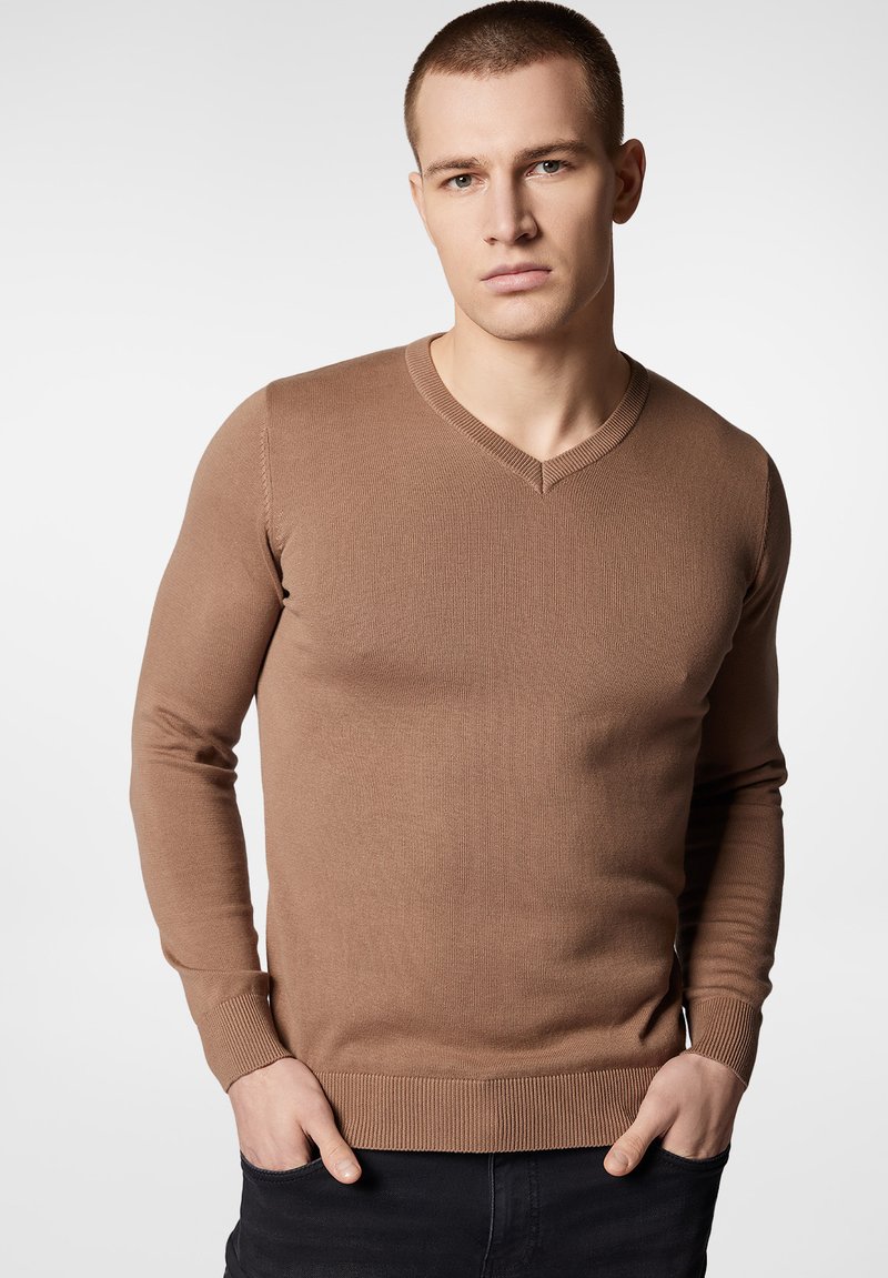Brązowy sweter z dekoltem w serek, z długimi rękawami, ribbowymi mankietami i dołem, dopasowany fason, wykonany z teksturowanej dzianiny, noszony z czarmi spodniami.