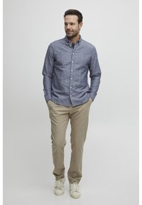 Camicia blu con bottoni, in tessuto testurizzato, maniche lunghe e un piccolo logo sul petto, abbinata a pantaloni chinos beige e sneakers bianche.