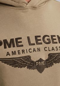 Beige hoodie met donkerbruine geborduurde tekst "PME LEGEND AMERICAN CLASSIC" en een gevleugeld embleem, gemaakt van een zachte, gestructureerde stof.