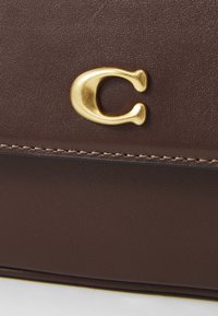 Braune Lederoberfläche mit feiner Naht und einem erhabenen goldenen Buchstaben "C"-Emblem, das zentriert in der Nähe der Naht platziert ist.