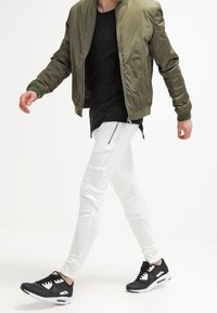 Pánský outfit zahrnující olivově zelenou bomber bundu, černé tričko, bílé joggers kalhoty s detaily na zip a černé sportovní boty s bílými akcenty.