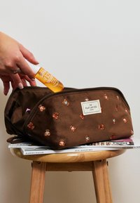 Ruskea kangasmeikkipussi, jossa on kukka-ompelukset, vetoketjukiinnitys ja logopatch. Käsi laittaa oranssia pulloa sisään.
