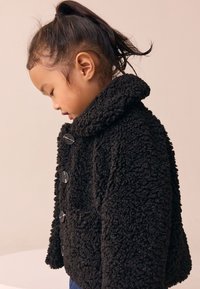 Black teddy coat with a soft, textured finish, featuring a folded collar, four large buttons, and a relaxed fit. Child shown side profile.