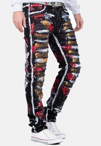 Jean noir en denim avec des détails distendus, éclaboussures de peinture rouge et or, et coupe slim. Présente des éléments en métal argenté et un accent ceinture cloutée.