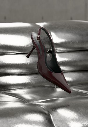 Scarpa slingback con tacco alto rosso scuro con punta affilata, appoggiata su una superficie imbottita argento metallizzato lucido.