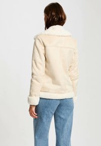 Veste en fausse fourrure beige clair avec un col et des poignets blancs duveteux, présentant une texture lisse et un design ajusté, vue de dos.