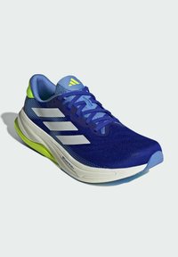 Blue athletic shoe with a textured mesh upper, white stripes, and a vibrant green heel accent. Features a cushioned sole and rounded toe.