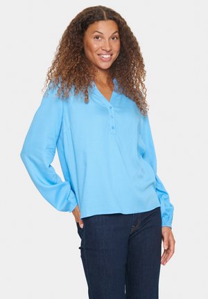 Blusa azzurra chiara con maniche lunghe e a sbuffo, scollatura a V e tre bottoni. Il tessuto ha un motivo testurizzato. Abbinata a jeans scuri.