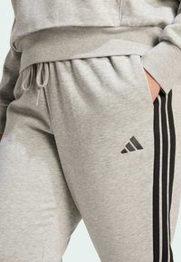 Harmaat collegehousut, jotka on valmistettu pehmeästä kankaasta, mustilla sivuraidoilla ja Adidaksen logolla vasemmassa reidessä, joustavan vyötärön kanssa.