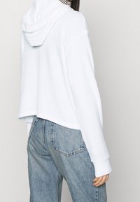 Sweat à capuche court blanc avec tissu texturé et grande capuche, associé à un jean denim bleu clair à coupe décontractée avec poches arrière.