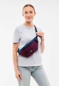 Riñonera color burdeos y azul marino con un bolsillo frontal con cremallera, correa ajustable y detalle del logo. Hecha de un tejido liso y duradero. Apropiada para un uso casual.