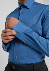 Camicia blu con bottoni, in tessuto strutturato, con bottoni marroni e maniche lunghe con polsini abbottonati, abbinata a pantaloni scuri.