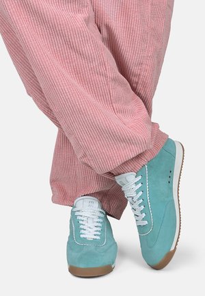 Persoon die roze corduroy broek en teal kleurige suède sneakers met witte veters draagt, staand met gekruiste benen tegen een witte achtergrond.