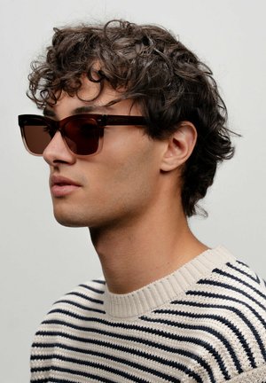 Lunettes de soleil rectangulaires avec des verres teintés marron, épaisses montures marron et détails texturés subtils. Portées avec un pull rayé.