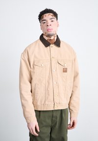Chaqueta de mezclilla beige con un cuello de pana marrón, que presenta bolsillos frontales, herrajes dorados y una etiqueta de cuero en el pecho.