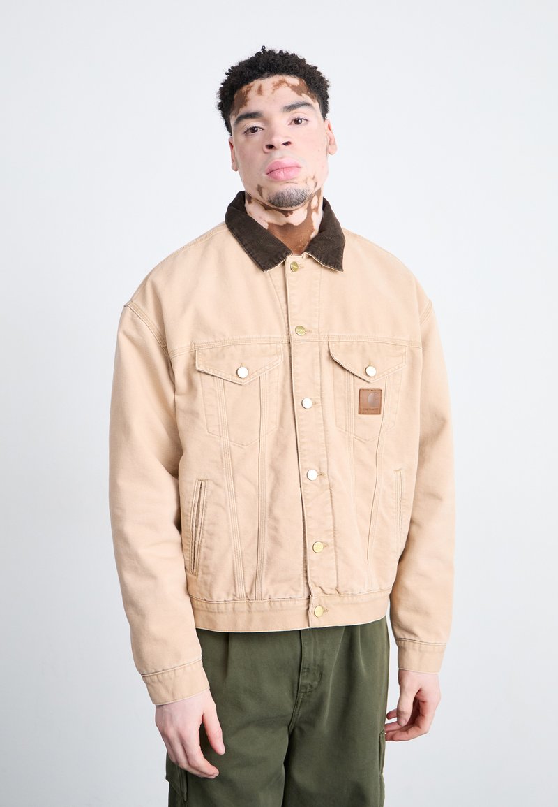 Chaqueta de mezclilla beige con un cuello de pana marrón, que presenta bolsillos frontales, herrajes dorados y una etiqueta de cuero en el pecho.