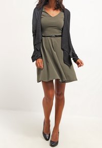 Žena v olivově zelených šatech s páskem, antracitově šedém kardiganu na zapínání a černých lodičkách stojí na bílém pozadí.
