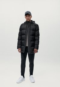 Melna pūkainā jaka ar kapuci, spīdīga tekstūra, priekšējie pogas, pārī ar melnām šaurām biksēm un baltām kedām. Tumša cepure noslēdz izskatu.