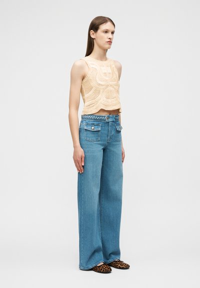 Femme portant un haut court en crochet beige sans manches, un jean bleu taille haute à jambes larges avec poches avant, et des chaussures à enfiler imprimé léopard.