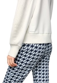 Weißes Sweatshirt mit strukturierten Strickärmeln und einem gerippten Saum, kombiniert mit blau-weiß gemusterten Leggings mit Logo-Details.