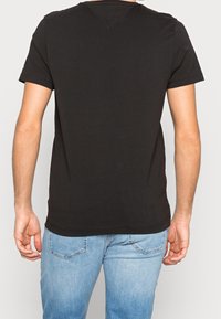Hombre con camiseta de manga corta negra sencilla y jeans azul claro, visto desde atrás contra un fondo liso claro.